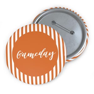 May include: A round button with an orange center and white vertical stripes around the edge. The center has the word "Gameday" in white script. The back of the button is silver with a pin.