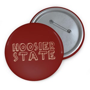 May include: Two round badges. The front badge is a deep red with the words "HOOSIER STATE" in a light orange, outlined font. The second badge is partially visible, showing a silver metal back with a pin.