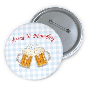 May include: A round button with a light blue and white checkered pattern. The button features two cartoon beer mugs clinking together, with the text "Cheers to gameday" in red script. The button has a silver pin back.