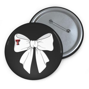 May include: Two round black buttons. One button features a white bow with a red and black "T" logo. The other button is silver with a pin back. The buttons are likely for clothing or accessories.