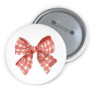 May include: A white button with a red and white gingham bow design. The bow is centered on the button and has a watercolor effect. The back of the button is silver with a pin.