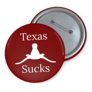 May include: A round, red button with the words "Texas Sucks" in white text. The button features a white longhorn silhouette. The back of the button is silver with a pin.