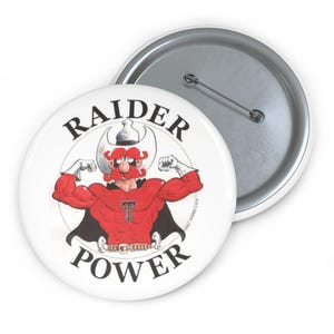 May include: White button with a cartoon illustration of a muscular figure in a red shirt and black cape. The text "RAIDER POWER" is printed around the figure. A second button is visible in the background.