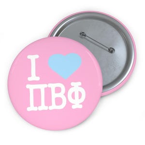 May include: A pink button with the text "I" and "ΠΒΦ" in white, and a light blue heart. The button has a silver pin back. The button is a round shape.