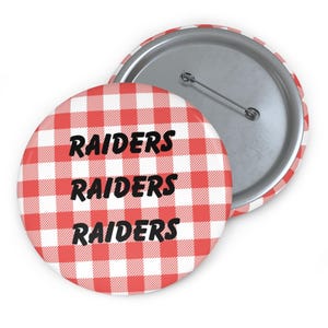 May include: Two round buttons with a red and white checkered pattern. The word "RAIDERS" is printed three times in black, stacked vertically. One button is face up, and the other is face down, showing the pin back.