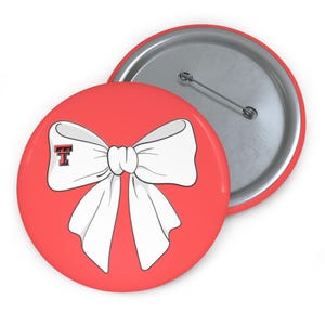 May include: A red button featuring a white bow design and a small red and black letter "T" logo. The button has a silver pin back. The bow is centered on the button, with the letter "T" in the upper left corner.