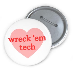 May include: A white button with a pink heart design. The heart contains the words "wreck 'em tech" in red lettering. The button has a silver pin backing.