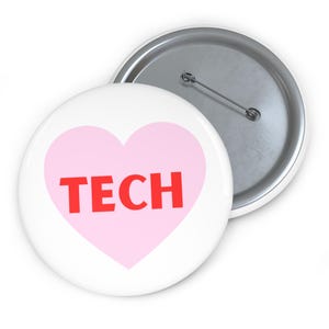 Pode incluir: Um botão branco com um design de coração rosa e a palavra "TECH" em vermelho. O botão tem um alfinete de metal na parte de trás e uma borda prateada. O design é simples e atraente.