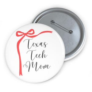 May include: White button with the text "Texas Tech Mom" in black script, accented by a red ribbon design. The button has a silver pin back.