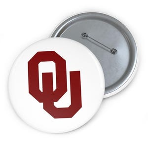 May include: Two white round buttons with a metal pin backing. One button features a maroon "OU" logo, the other button is partially visible. The buttons are likely for clothing or accessories.