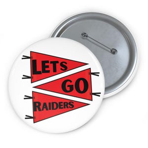 May include: A white button with a metal backing. The button features the text "LET'S GO RAIDERS" in black letters on two red pennant shapes. The button is a fan accessory.