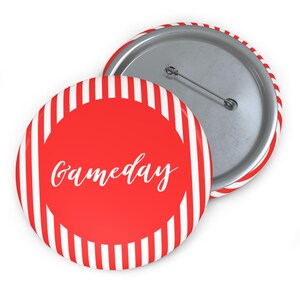 May include: A round button with a red center and the word "Gameday" in white script. The button has a red and white striped border and a metal pin back. The button is a fun accessory for game day.