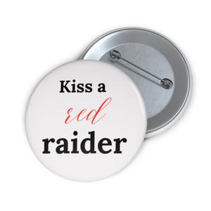 May include: White button with the text "Kiss a red raider" in black and red. The button has a metal pin back. The button is round and has a glossy finish.