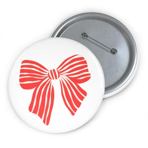 May include: Two white button pins. One pin features a red bow design with white stripes. The other pin is silver with a pin back. The bow design is centered on the white button.