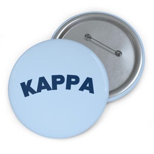 May include: Two round buttons. One button is light blue with the word "KAPPA" in dark blue. The other button is silver with a pin back. The buttons are on a white surface.