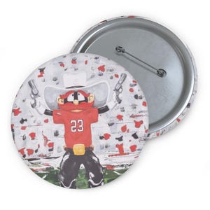 May include: A round button with a cartoon cowboy design. The cowboy wears a red shirt with the number 23, a white hat, and holds two guns. The background is white with red and black paint splatters. The button has a metal pin back.
