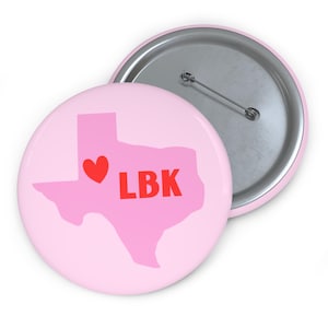 May include: A light pink button with a pink Texas outline, a red heart, and the letters "LBK" in red. The button has a silver pin backing and a silver rim.