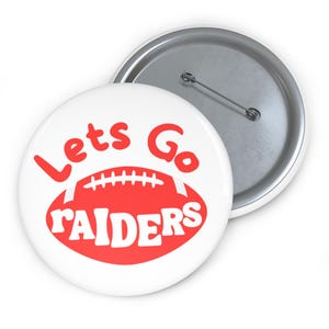May include: White button with a red football graphic and the words "Lets Go RAIDERS" in red. The button has a silver pin back and is a sports-themed accessory.