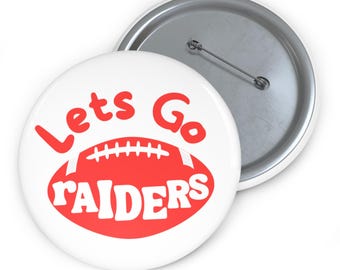 Puede incluir: Botón blanco con un gráfico de fútbol americano rojo y las palabras "Lets Go RAIDERS" en rojo. El botón tiene un alfiler plateado en la parte posterior y es un accesorio deportivo.