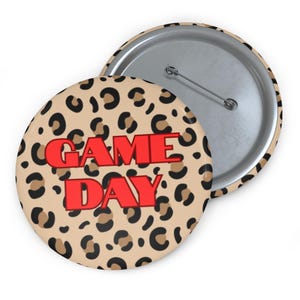 May include: A round button with a tan leopard print background and the words "GAME DAY" in red block letters. The button has a metal pin back and a silver rim.