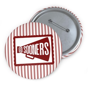 May include: Two round buttons with a red and white striped border. One button features a red megaphone design with the words "GO SOONERS" in white. The other button has a silver pin back.
