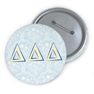 May include: A round button with a light blue floral pattern background. Three yellow and blue outlined triangles are centered on the button. The button has a metal pin back.