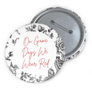 May include: A white button with a floral design border and the text "On Game Days We Wear Red" in red. The button has a metal pin back and a silver rim.