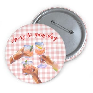 May include: A round button with a pink and white gingham pattern. The button features an illustration of three hands holding cocktails. The text "Cheers to gameday" is written in red script. The button has a silver pin back.