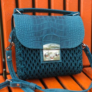 May include: Teal crossbody bag with a textured flap and a woven body. The bag features a silver clasp and a matching adjustable strap. The bag is sitting on an orange surface.