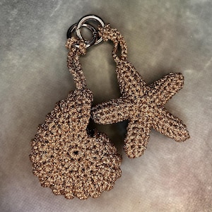 Puede incluir: Un par de llaveros de ganchillo de color bronce. Uno tiene forma de concha y el otro de estrella de mar. Ambos están unidos a un anillo de metal plateado.