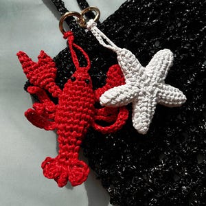 以下が含まれることがあります： 黒いかぎ針編みのトートバッグ。金色のリングに赤いかぎ針編みのロブスターと白いかぎ針編みのヒトデが付いています。バッグには黒いハンドルとテクスチャード加工が施されています。ロブスターとヒトデは、目に見えるステッチで詳細に表現されています。