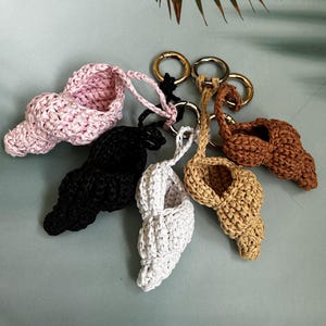 May include: A collection of six handmade seashell-shaped keychains in various colors: pink, black, white, tan, and brown. Each keychain features a woven texture and a gold or silver-colored metal ring for attaching keys. The keychains are arranged on a light blue surface.