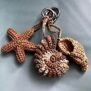 May include: Three woven raffia keychains in the shape of a starfish, a seashell, and a spiral shell. The keychains are attached to silver metal rings. The raffia is in shades of brown and beige.