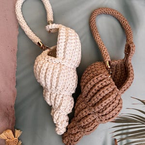 May include: Two crochet seashell-shaped handbags. One is off-white, the other is tan. Both have a thick, rope-like handle attached with gold-colored hardware. The bags are on a light blue surface with decorative elements.