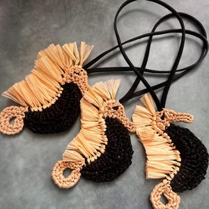 May include: Three seahorse-shaped ornaments crafted from black crocheted yarn and light beige raffia-like fringe. Each seahorse features a curved body, a textured mane, and a coiled tail, attached to a thin black cord for hanging or display purposes.