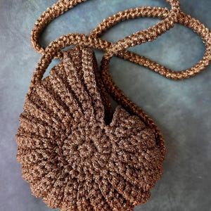 Può includere: Una borsa all'uncinetto color bronzo metallizzato a forma di conchiglia di nautilo. La borsa presenta un motivo a spirale testurizzato realizzato con filato scintillante e include una lunga tracolla intrecciata abbinata per il trasporto.