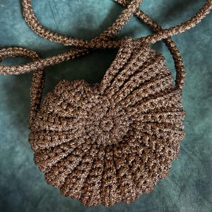 Op de afbeelding: Een handgemaakte, ronde, goudkleurige gehaakte tas met een lange riem. De tas heeft een geschulpte rand en een spiraalvormig ontwerp dat vanuit het midden uitstraalt. De riem is ook gehaakt en lijkt stevig.