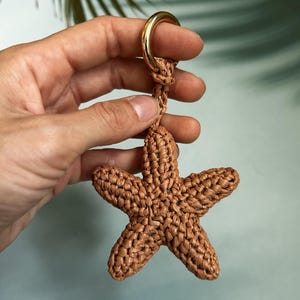 May include: A brown, five-pointed starfish-shaped keychain crafted from woven cord. The starfish is attached to a polished gold-tone metal ring, providing a secure loop for keys or accessories. The texture is consistent and knotted throughout.