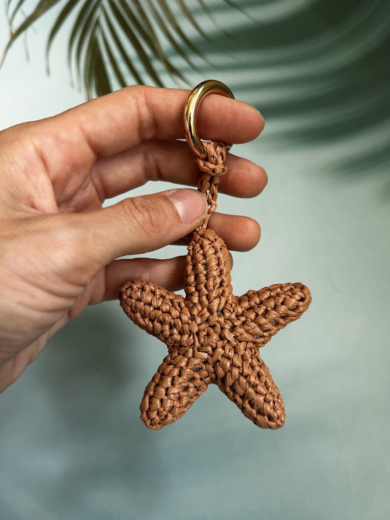 May include: A brown, five-pointed starfish-shaped keychain crafted from woven cord. The starfish is attached to a polished gold-tone metal ring, providing a secure loop for keys or accessories. The texture is consistent and knotted throughout.