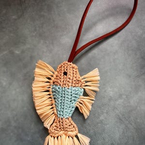 May include: A fish-shaped macrame ornament crafted from tan and light blue cord, featuring fringed fins and a tail made of light tan raffia-like material, suspended by a thin, dark red leather loop for hanging.