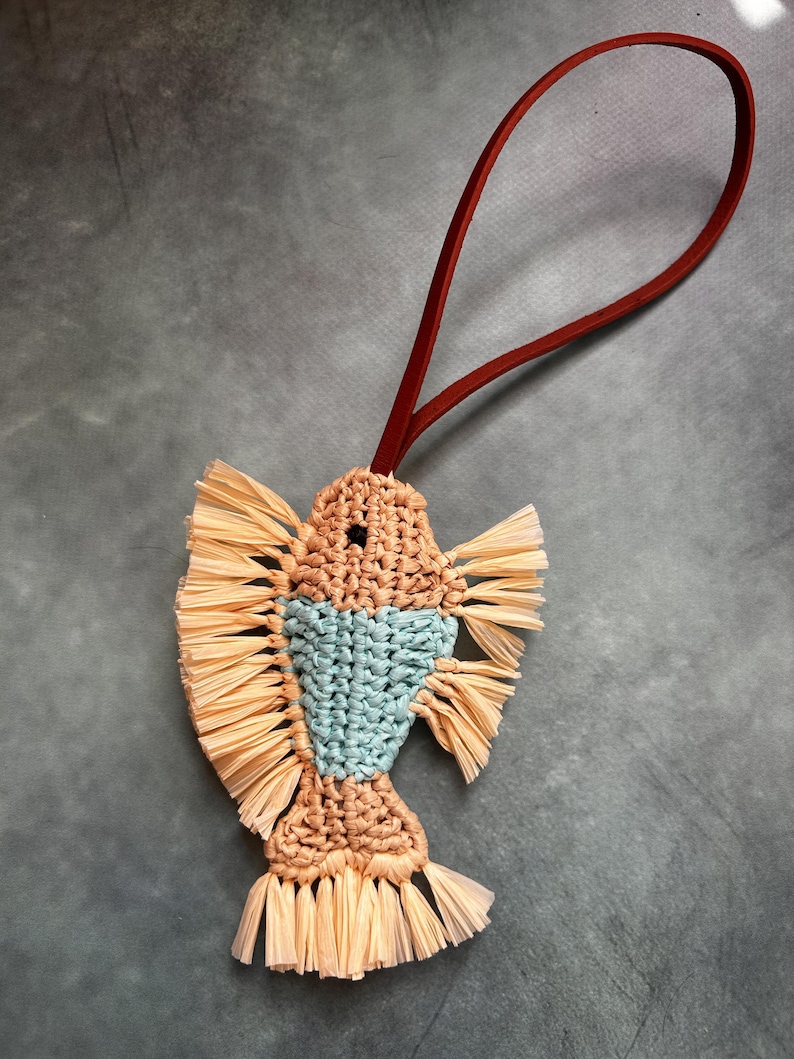May include: A fish-shaped macrame ornament crafted from tan and light blue cord, featuring fringed fins and a tail made of light tan raffia-like material, suspended by a thin, dark red leather loop for hanging.