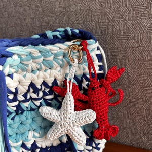 May include: A square crochet bag featuring a striped pattern of dark blue, light blue, and white yarn. Attached to a gold-toned metal ring on the bag are two crochet charms: a white five-pointed starfish and a bright red lobster with black eyes.