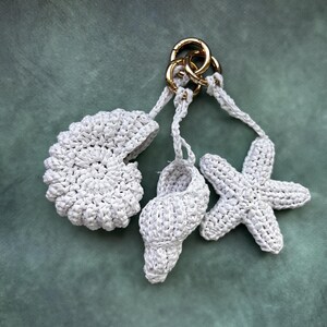 Puede incluir: Un llavero blanco de ganchillo con una concha, una caracola y una estrella de mar. Los amuletos est&aacute;n unidos a un cierre dorado. El fondo es de color verde azulado apagado.