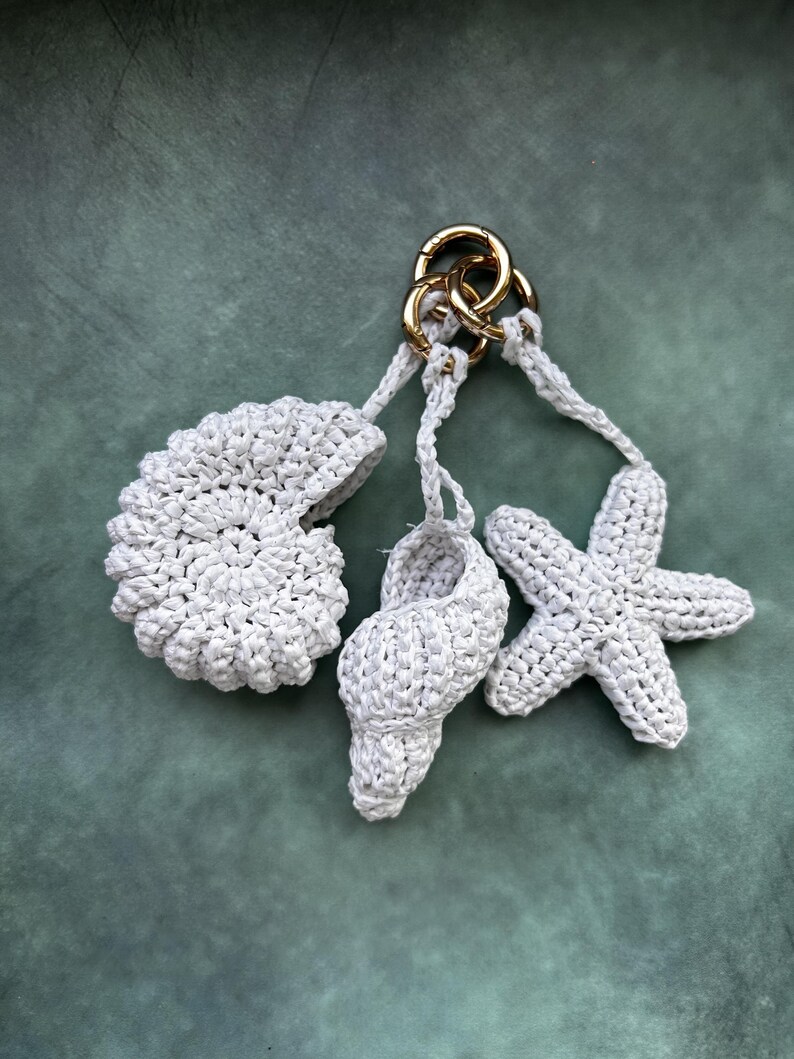 Puede incluir: Un llavero blanco de ganchillo con una concha, una caracola y una estrella de mar. Los amuletos est&aacute;n unidos a un cierre dorado. El fondo es de color verde azulado apagado.