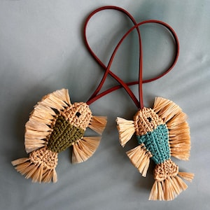 May include: Two fish-shaped ornaments crafted from woven cord and raffia-like fringe. One fish features a dark olive green body, the other a teal body, both with tan heads and tails. Each is attached to a thin, dark red leather cord loop.