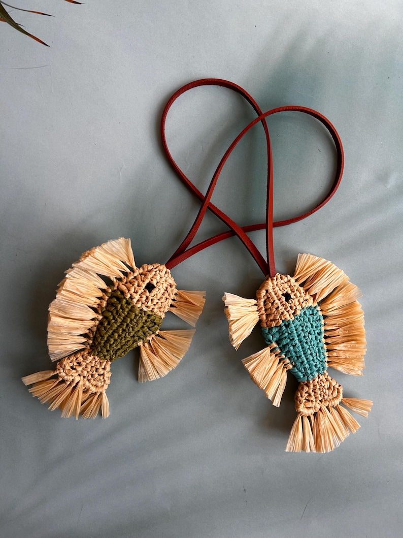 May include: Two fish-shaped ornaments crafted from woven cord and raffia-like fringe. One fish features a dark olive green body, the other a teal body, both with tan heads and tails. Each is attached to a thin, dark red leather cord loop.