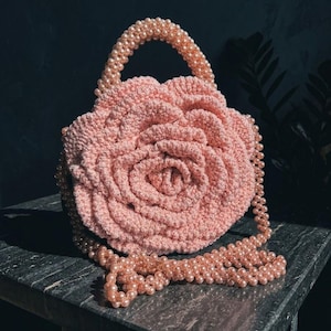 May include: A light pink crochet handbag shaped like a large blooming rose. The bag features a structured handle and a long shoulder strap, both entirely covered in small, round, light pink faux pearls that match the color of the yarn.