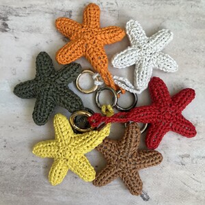 May include: A collection of six crocheted starfish keychains in orange, white, dark green, red, yellow, and brown. Each starfish is crafted with a textured yarn stitch and features a circular metal ring attached to one of its five arms.