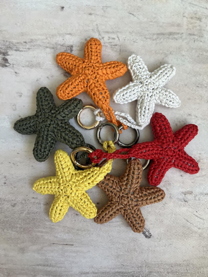 May include: A collection of six crocheted starfish keychains in orange, white, dark green, red, yellow, and brown. Each starfish is crafted with a textured yarn stitch and features a circular metal ring attached to one of its five arms.