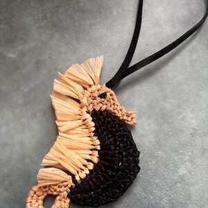 May include: A seahorse-shaped ornament crafted from woven raffia in natural beige and black. The seahorse features a fringed beige dorsal fin, a solid black body, and a curled tail. A thin black cord is attached to the top for hanging purposes.